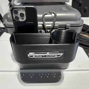 Pode incluir: Um organizador de plástico preto com um suporte para telefone, um porta-copos e um rótulo que diz "East Cape".