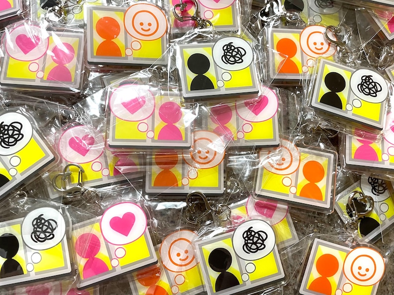 May include: A collection of colorful keychains with different designs. Each keychain features a small square with two images inside. The images include a heart, a smiley face, and a tangled line. The keychains are packaged in clear plastic bags.