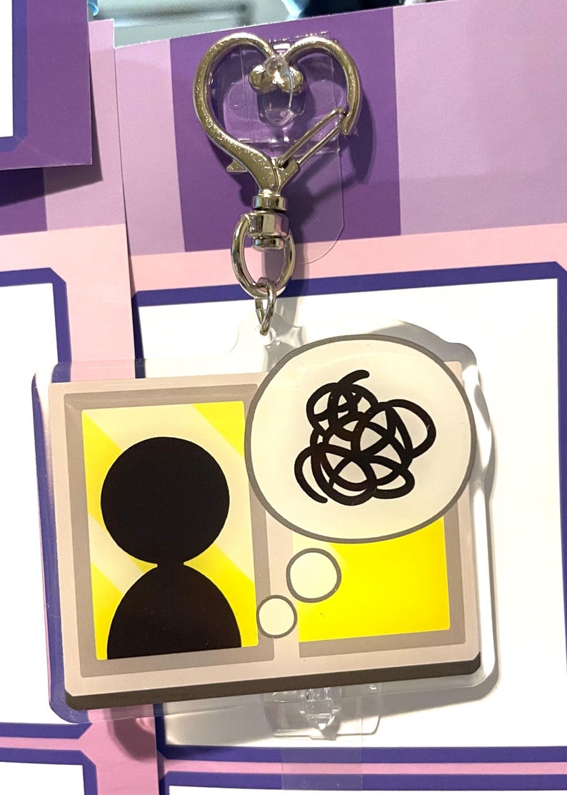 May include: A clear acrylic keychain with a silver heart-shaped clasp. The keychain features a cartoon illustration of a person with a thought bubble filled with a black scribble.