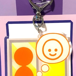 May include: A white and yellow keychain with a silver clip. The keychain features a cartoon person with a thought bubble above their head. The thought bubble contains a smiling face.