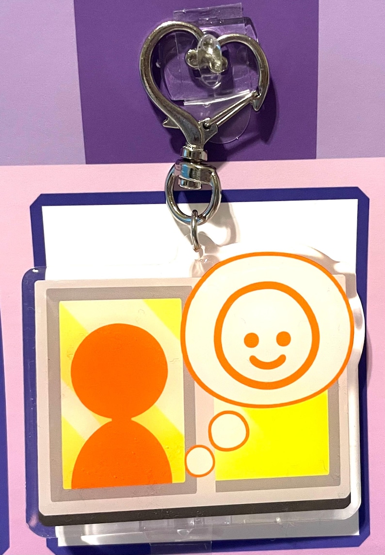 May include: A white and yellow keychain with a silver clip. The keychain features a cartoon person with a thought bubble above their head. The thought bubble contains a smiling face.