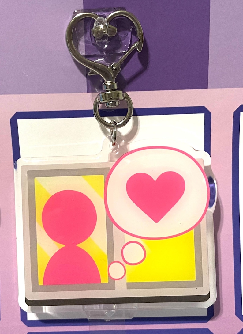 May include: A pink and white acrylic keychain with a cartoon person in a window with a thought bubble containing a pink heart. The keychain has a silver heart-shaped clasp.