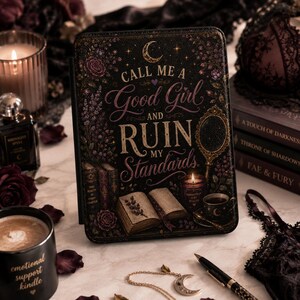 Puede incluir: Una funda para lector electrónico negra con una ilustración dorada y morada de un libro abierto, una vela, un espejo de mano y flores. El texto dice: CALL ME A Good Girl AND RUIN MY Standards. El diseño incluye una luna creciente y estrellas.