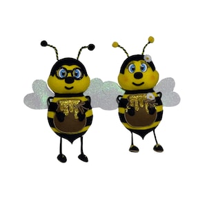May include: Two felt bee figurines with black and yellow stripes, holding honey pots. One bee is wearing glasses and the other has a flower on its head.