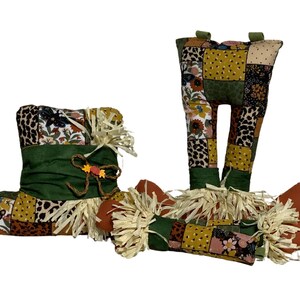 May include: A patchwork fabric scarecrow with a green hat, brown legs, and straw accents. The fabric features a floral and animal print pattern in shades of green, yellow, brown, and pink.