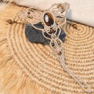 Peut inclure: Un ornement de main en macramé beige avec une pierre précieuse centrale en œil-de-tigre ovale. Le design complexe en cordon noué est agrémenté de petites perles métalliques et d'autres pierres œil-de-tigre, formant un motif symétrique avec une boucle.