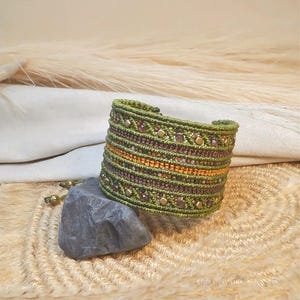 Può includere: Un bracciale a fascia largo realizzato in corda verde oliva, caratterizzato da un complesso intreccio in macramè, file orizzontali di filo marrone scuro, una banda centrale arancione metallizzato e perline color bronzo e cristalli sfaccettati.