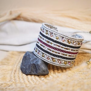 May include: A wide woven cuff bracelet featuring horizontal bands of beige, black, and deep burgundy thread. The design is accented with rows of small metallic gold beads and faceted light pink crystals integrated into the intricate macrame pattern.