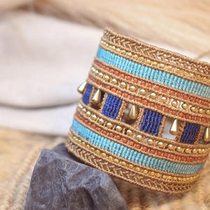 Könnte beinhalten: Ein breites Makramee-Armband mit horizontalen Bändern aus hellblauem, dunkelblauem und orangefarbenem Garn. Das Design ist mit Reihen kleiner goldfarbener Perlen und hängenden, tropfenförmigen Anhängern in Goldoptik im Gewebe verziert.