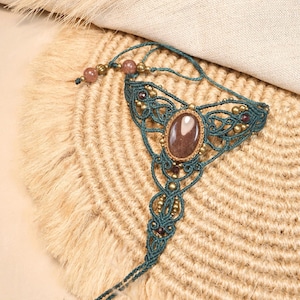 Pode incluir: Um ornamento de mão em macramé na cor verde-azulado com uma pedra castanha oval central, padrões de cordão atado intrincados e pequenas contas metálicas. O design inclui uma pulseira ligada a um laço para o dedo com contas metálicas.