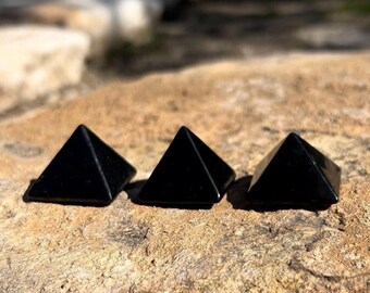 May include: Three black obsidian pyramids on a rough, brown rock surface. The pyramids are smooth and polished.