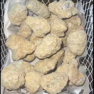 May include: A collection of rough, light brown and gray stones with a slightly textured surface. The stones are arranged in a white wire basket lined with white paper.