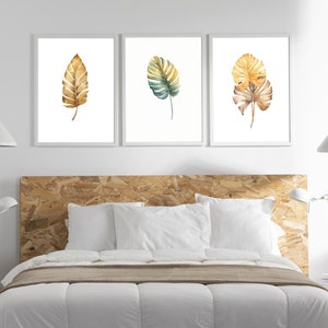 Könnte beinhalten: Drei gerahmte Aquarelldrucke von tropischen Blättern in Gold- und Grüntönen. Die Drucke hängen an einer weißen Wand über einem Bett mit einem Holz-Kopfteil.