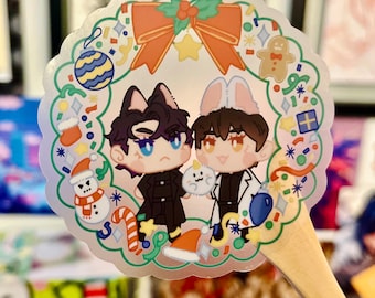 May include: A clear sticker with a Christmas wreath design featuring two cartoon characters, one with black cat ears and the other with bunny ears. The characters are wearing winter clothing and holding a small white animal. The sticker is on a wooden stick.