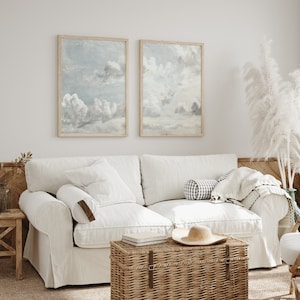 Könnte beinhalten: Ein weißes Leinen-Sofa mit zwei Kissen, einer Wolldecke und einer Aufbewahrungstruhe aus Weide. Zwei gerahmte Drucke eines bewölkten Himmels hängen an der Wand hinter dem Sofa.