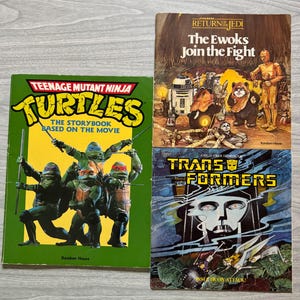 May include: Three vintage children's books are displayed. The books include "Teenage Mutant Ninja Turtles," "Return of the Jedi: The Ewoks Join the Fight," and "Transformers." The covers feature colorful illustrations and text, with the "Teenage Mutant Ninja Turtles" book in green.