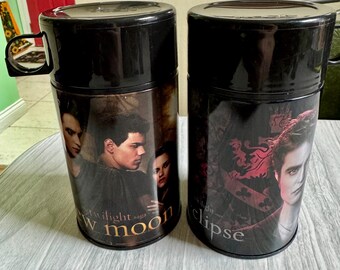 May include: Two black travel mugs featuring images from the Twilight saga. One mug displays the text "Twilight saga New Moon" with character portraits. The other mug has "Twilight Eclipse" with a character portrait. Both mugs have black lids and handles.