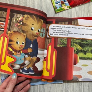 K&ouml;nnte beinhalten: Eine Illustration aus einem Kinderbuch mit Daniel Tiger und seinem Vater in einer Stra&szlig;enbahn. Die Stra&szlig;enbahn ist rot und gelb. Der Text im Bild lautet: "Bitte bring uns zur Schule, Trolley." Das Buch ist auf einem Tisch ge&ouml;ffnet.