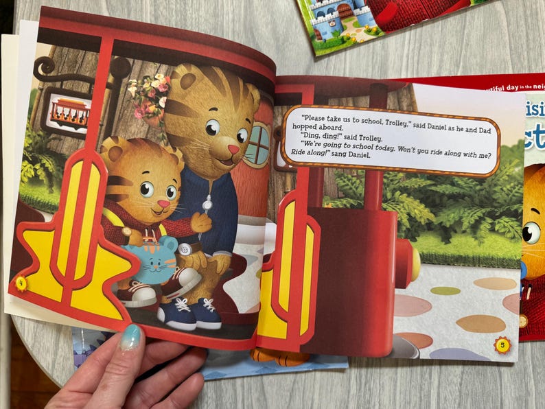 K&ouml;nnte beinhalten: Eine Illustration aus einem Kinderbuch mit Daniel Tiger und seinem Vater in einer Stra&szlig;enbahn. Die Stra&szlig;enbahn ist rot und gelb. Der Text im Bild lautet: "Bitte bring uns zur Schule, Trolley." Das Buch ist auf einem Tisch ge&ouml;ffnet.