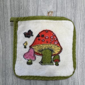 May include: A square white potholder with a green fabric border and a central tufted design featuring a large red mushroom with pink spots, a smaller pink mushroom, a tiny green mushroom, and a small dark butterfly above the mushrooms.