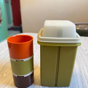 May include: A stack of three vintage plastic containers in orange, olive green, and brown. Next to it is a rectangular olive green container with a white lid. The containers are on a light gray surface.