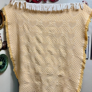 May include: A yellow blanket with a heart pattern and white fringe. The blanket is made of a textured material and has a decorative design. The blanket is a home decor item.