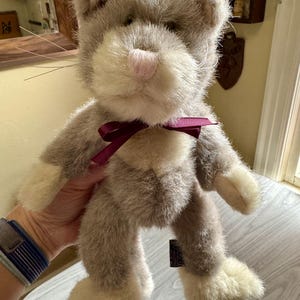May include: A plush cat toy with grey and cream-colored faux fur, featuring a soft pink nose, long thin whiskers, and a dark purple ribbon tied around its neck. The stuffed animal has cream-colored paws and a small black fabric tag on its side.