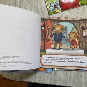 K&ouml;nnte beinhalten: Ein aufgeschlagenes Kinderbuch mit einer farbenfrohen Illustration von Daniel Tiger und seinem Vater. Der Text lautet: &bdquo;Hallo Nachbar! Es ist Zeit f&uuml;r die Schule!&ldquo; Weitere B&uuml;cher sind im Hintergrund zu sehen.