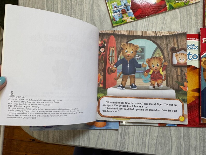 K&ouml;nnte beinhalten: Ein aufgeschlagenes Kinderbuch mit einer farbenfrohen Illustration von Daniel Tiger und seinem Vater. Der Text lautet: &bdquo;Hallo Nachbar! Es ist Zeit f&uuml;r die Schule!&ldquo; Weitere B&uuml;cher sind im Hintergrund zu sehen.
