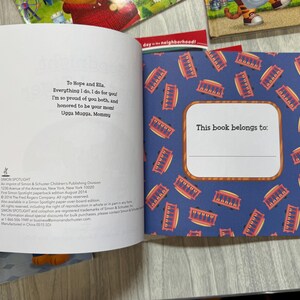 K&ouml;nnte beinhalten: Aufgeschlagenes Buch mit blauem Einband und einem Muster aus roten und goldenen Stra&szlig;enbahnen. Die Innenseiten enthalten eine Widmung und einen Bereich mit der Aufschrift "This book belongs to:" zur Personalisierung. Das Buch geh&ouml;rt zur Simon Spotlight-Reihe.