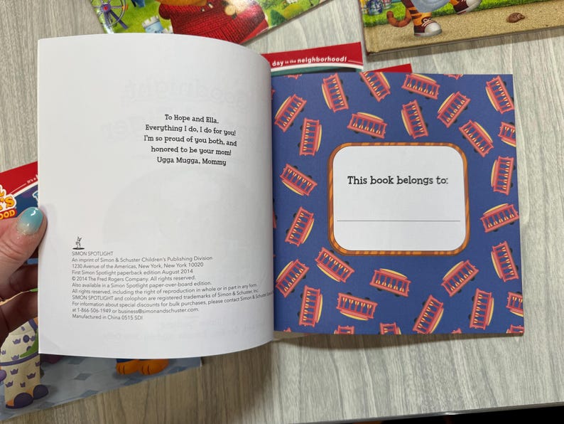 K&ouml;nnte beinhalten: Aufgeschlagenes Buch mit blauem Einband und einem Muster aus roten und goldenen Stra&szlig;enbahnen. Die Innenseiten enthalten eine Widmung und einen Bereich mit der Aufschrift "This book belongs to:" zur Personalisierung. Das Buch geh&ouml;rt zur Simon Spotlight-Reihe.
