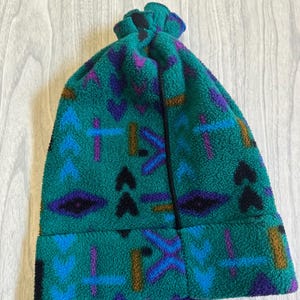 May include: A teal fleece beanie featuring a repeating geometric pattern in shades of purple, light blue, black, and brown. The hat has a cuffed brim and a gathered top, constructed from a soft, textured fabric with a visible vertical seam.