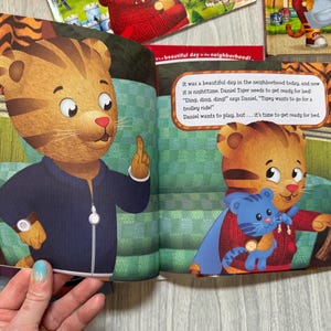 K&ouml;nnte beinhalten: Eine Kinderbuchillustration mit Daniel Tiger, einer animierten Tigerfigur, in einer marineblauen Jacke. Der Text lautet: &bdquo;Es war heute ein wundersch&ouml;ner Tag in der Nachbarschaft, und jetzt ist es Nacht. Daniel Tiger muss sich fertig machen, um ins Bett zu gehen!"