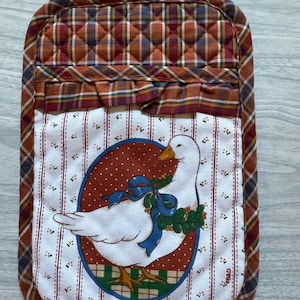 May include: A quilted oven mitt with a plaid pattern in shades of brown, red, and orange. The center features a white goose illustration within a red dotted oval, accented with a blue bow and green wreath. A loop is attached for hanging.