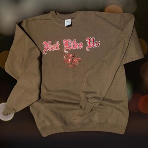 May include: A brown sweatshirt with the text "Not Like Us" in red gothic lettering and a red logo. The crew neck sweatshirt has long sleeves and ribbed cuffs and hem.