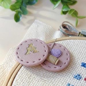 May include: Two pink, round, decorative sewing accessories. One has a gold letter "A", the other has a needle, thread, and a small purple heart. The accessories are on a white cross-stitch fabric.