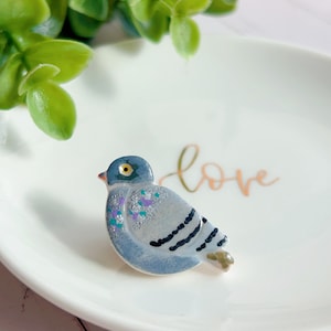 Può includere: Una statuetta di colomba in ceramica nei toni del blu, grigio e argento, con un tocco di viola e verde acqua. L'uccello è posato su un piatto bianco con la parola "love" scritta in corsivo. Foglie verdi sullo sfondo.