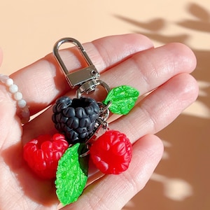 May include: A silver keyring with a cluster of colourful berries. The keyring includes a black blackberry, two red raspberries, and green leaves. The berries have a textured surface, and the leaves have a detailed, veined appearance.