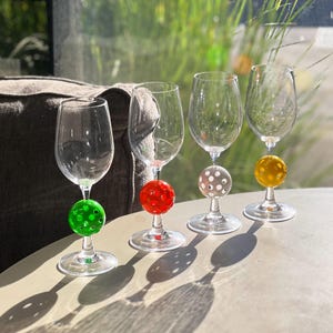 May include: Four clear wine glasses with different colored balls attached to the stems. The balls are red, green, white, and yellow.