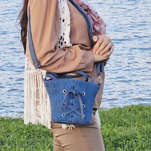 Könnte beinhalten: Eine blaue Jeans-Eimer-Tasche mit Schnürverschluss und langem Trageriemen. Die Tasche wird von einer Person getragen, die ein braunes Hemd und eine Hose trägt.