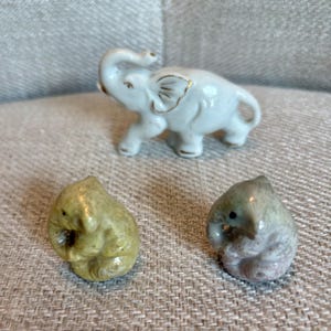 May include: A white ceramic elephant figurine with gold-painted ear accents positioned behind two smaller, rounded stone animal carvings, one in a muted yellow-green tone and the other in a mottled grey-brown hue, all featuring smooth surfaces.