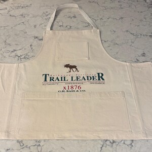 Peut inclure: Un tablier en toile blanc cassé avec un motif de wapiti marron et le texte Backwoods Territory TRAIL LEADER AUTHORITY EXPERIENCE GUIDANCE x1876 G.H. BASS & CO. Le tablier comporte une petite poche sur la poitrine et une grande poche frontale.