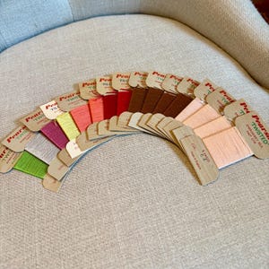 May include: A collection of Pearsall's Twisted Embroidery Silk thread cards arranged in a fan shape. The cards feature a spectrum of colors including green, silver, magenta, yellow, coral, red, brown, and peach, each labeled 8 yards.