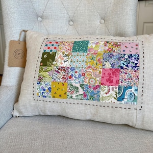 May include: Rectangular linen pillow with a patchwork design. The patchwork features small, colorful floral and patterned fabric squares. The pillow has a beige background with a black stitched border. A brown tag hangs from the pillow.