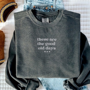 May include: A dark gray sweatshirt with the text "these are the good old days" in white lettering. The sweatshirt is paired with blue jeans and brown platform shoes.
