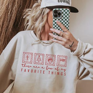 Puede incluir: Una sudadera color crema con la frase roja "These are a few of my favorite things" y cuatro gráficos estilo sello postal. La persona lleva una gorra de béisbol blanca con "Malibu Cowboy" y sostiene una funda de teléfono a cuadros verdes y blancos.