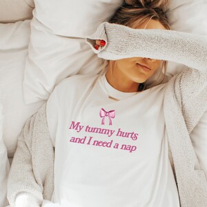 May include: White t-shirt with pink text that reads "My tummy hurts and I need a nap" and a pink bow graphic.