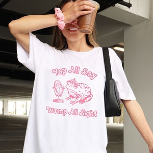 Puede incluir: Camiseta blanca de gran tamaño con un gráfico rosa de una rana con una corona que se mira en un espejo. El texto "YAP ALL DAY WOMP ALL NIGHT" está impreso debajo de la rana.
