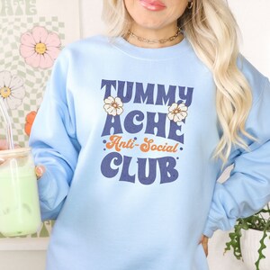 May include: A light blue sweatshirt with the text "Tummy Ache Anti-Social Club" in a retro font. The text is in navy blue with orange accents. There are two small white flowers on either side of the text.