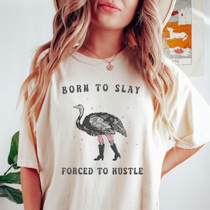 Peut inclure: Un t-shirt blanc avec une illustration noir et blanc d'une autruche portant des nœuds roses et des bottes noires. Le texte "Born to Slay" et "Forced to Hustle" est imprimé sur le t-shirt.